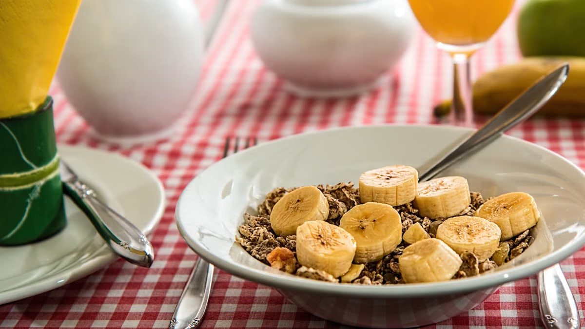 A healthy breakfast bowl with sliced bananas and cereal on a red checkered tablecloth.