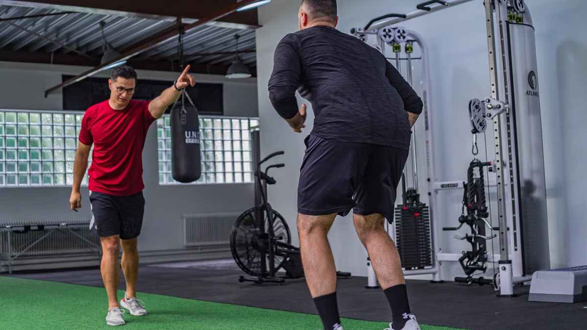 Back view of muscular man in sportswear doing exercises with trainer in modern fitness club